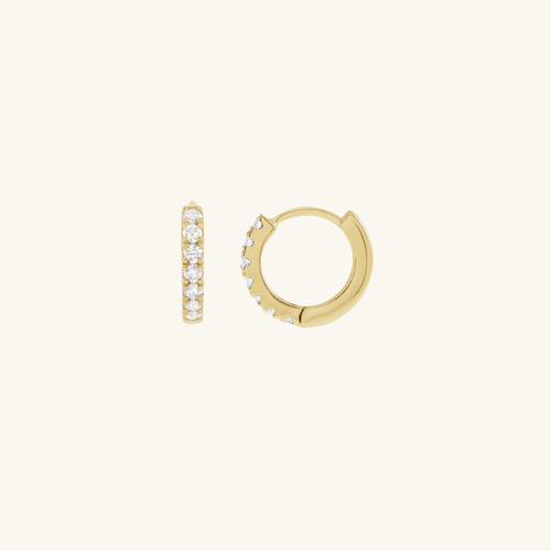A pair of gold huggie hoop earrings, one shown from the front with a vertical row of clear pavé-set stones and the other from the side showing the hinge and the stone-set edge, on a solid off-white background.